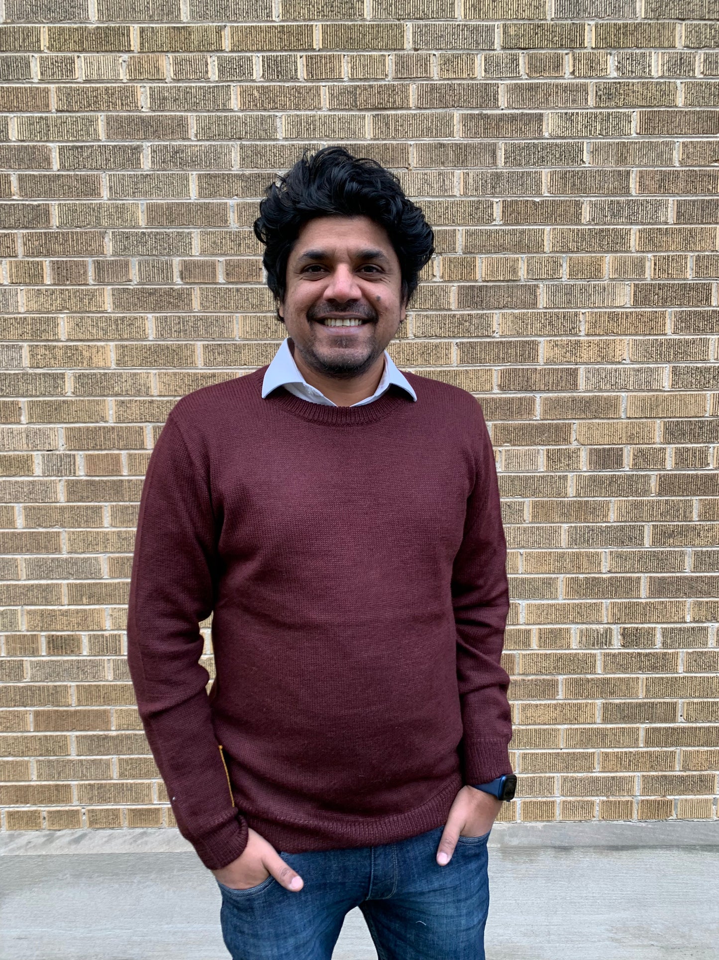 Man wearing a maroon sweater and blue jeans standing in front of a brick wall.