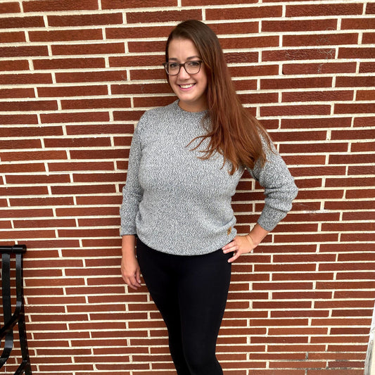 Woman wearing a gray sweater and black pants standing against a brick wall.
