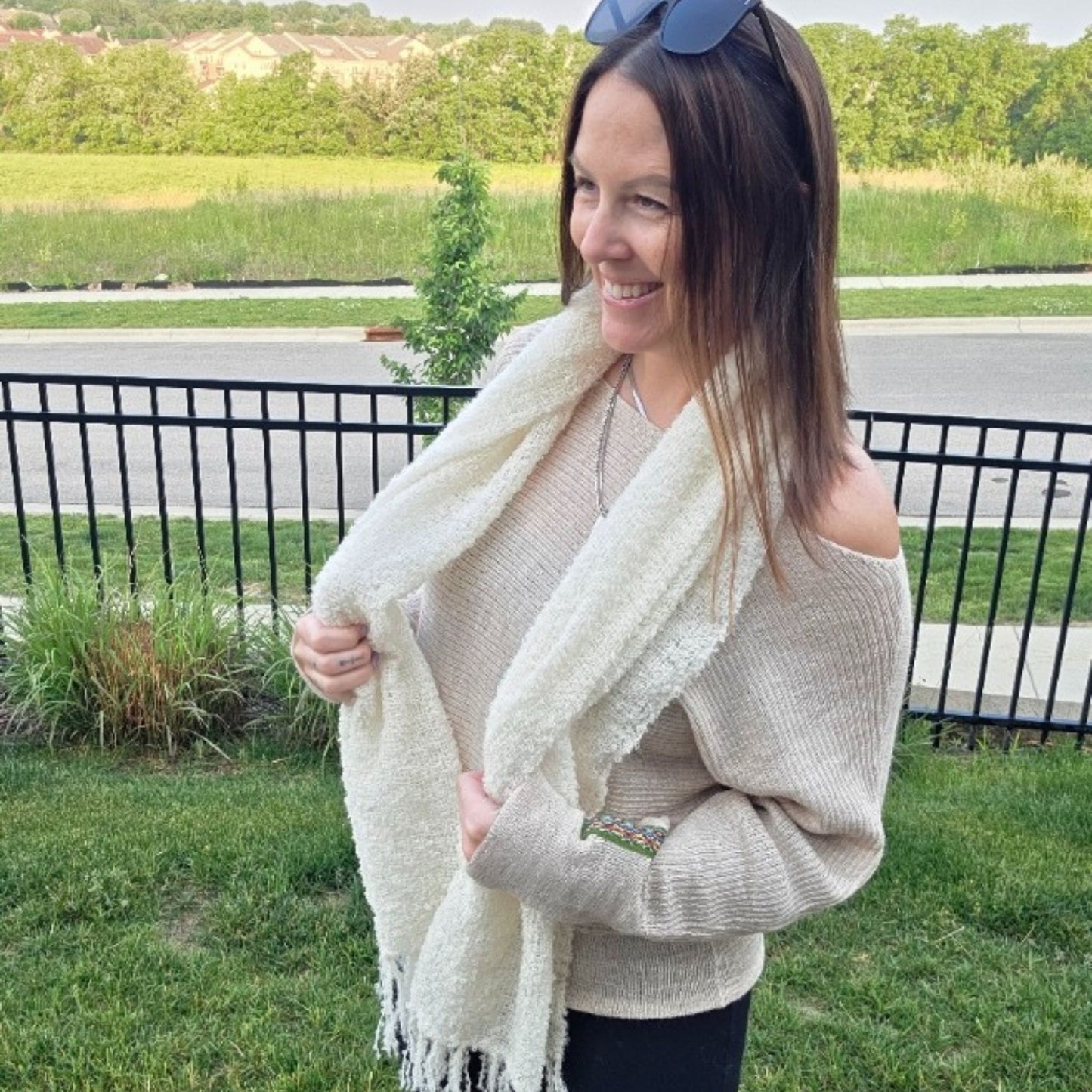 Woman wearing a beige sweater and white scarf outdoors with greenery in the background