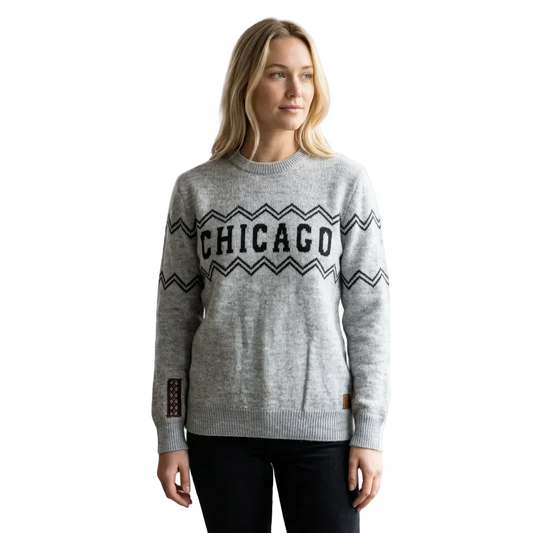 A woman modeling a light gray knit crewneck sweater with 'CHICAGO' in black lettering across the chest, framed by black zigzag patterns, against a solid beige background.