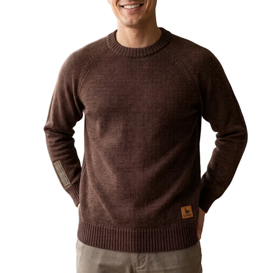 A man wearing a dark brown ribbed knit crewneck sweater with a small brown leather logo patch on the lower hem and a textured sleeve patch, set against a solid beige background.