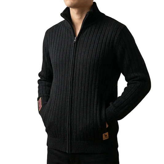 A man wearing a black ribbed knit zip-up sweater with a stand collar, side pockets, and small brown brand patches on the sleeve and hem, shown against a plain beige background.