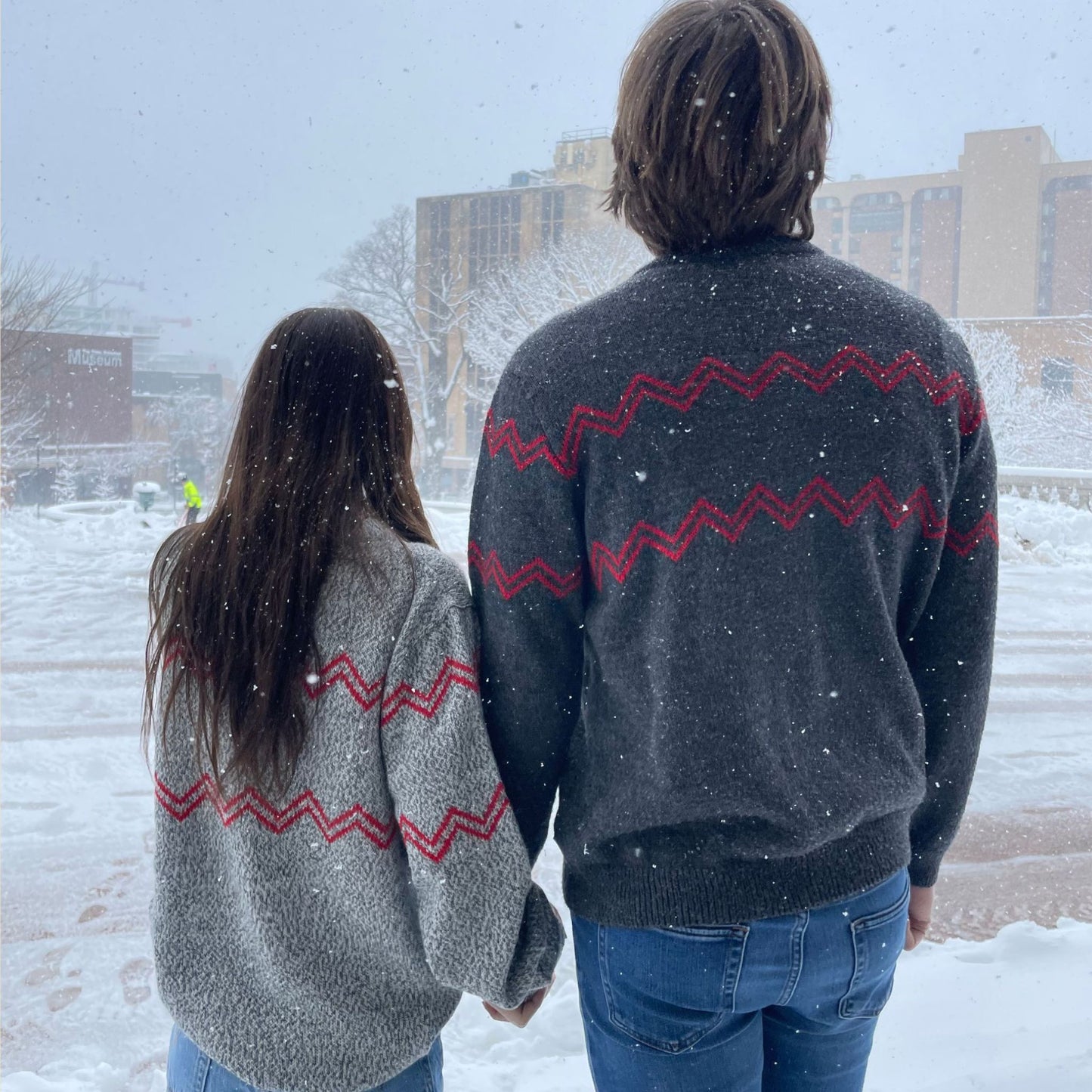 Two people wearing matching gray sweaters with red zigzag patterns standing in a snowy outdoor setting.