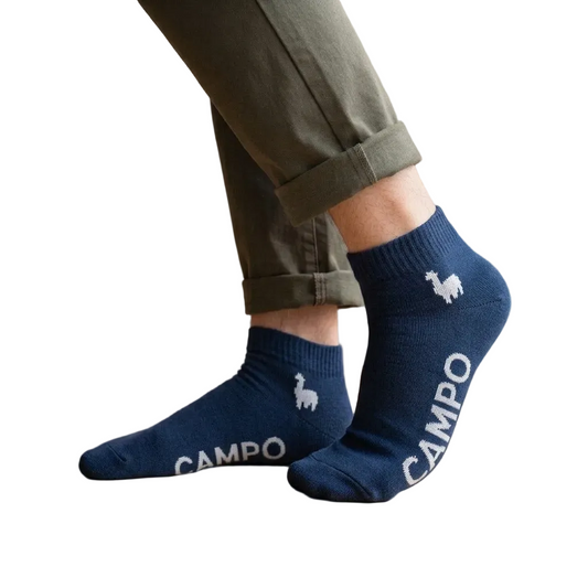 A person wearing navy blue ankle socks with white llama graphics and the brand name 'CAMPO' printed on the side, paired with olive green pants in a bright indoor setting with a wooden floor and window.