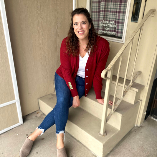 Woman in a red cardigan and blue jeans sitting on steps outside a building.