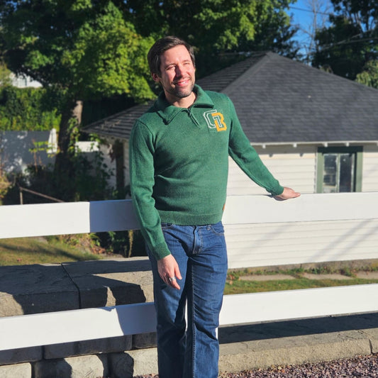 Man wearing a green sweater with a logo, standing outdoors by a white fence.