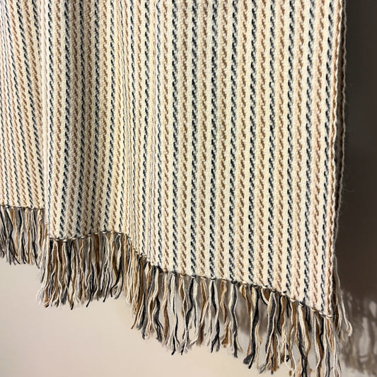 A close-up of a woven throw blanket with a cream, beige, and dark gray striped pattern and fringed edges, draped against a neutral background.