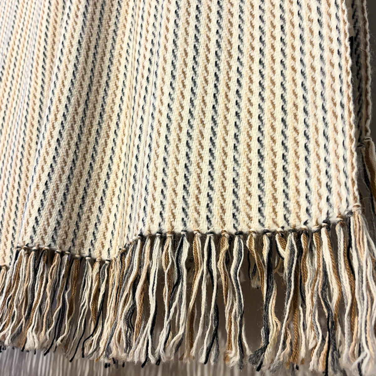 Close-up of a woven throw blanket with a cream, beige, and dark gray striped pattern and fringed edges.