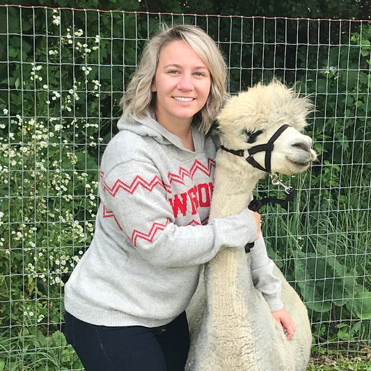 Woman in a gray hoodie with red text standing next to a white alpaca in an outdoor setting.