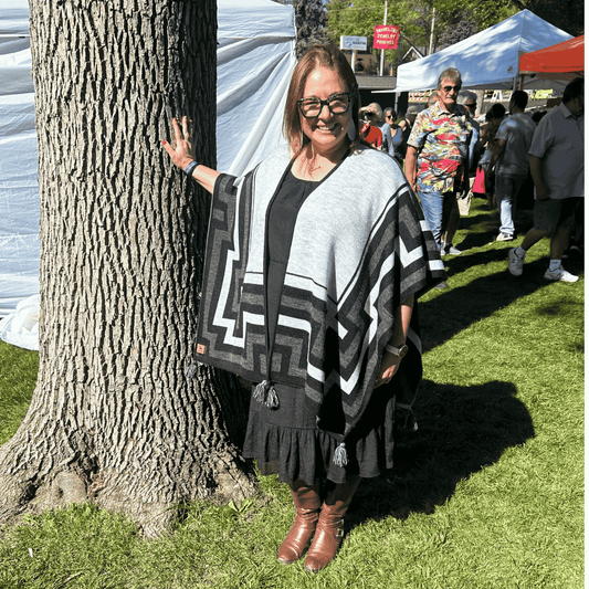 Woman wearing a patterned  Alpaca shawl leaning against a tree at an outdoor event.