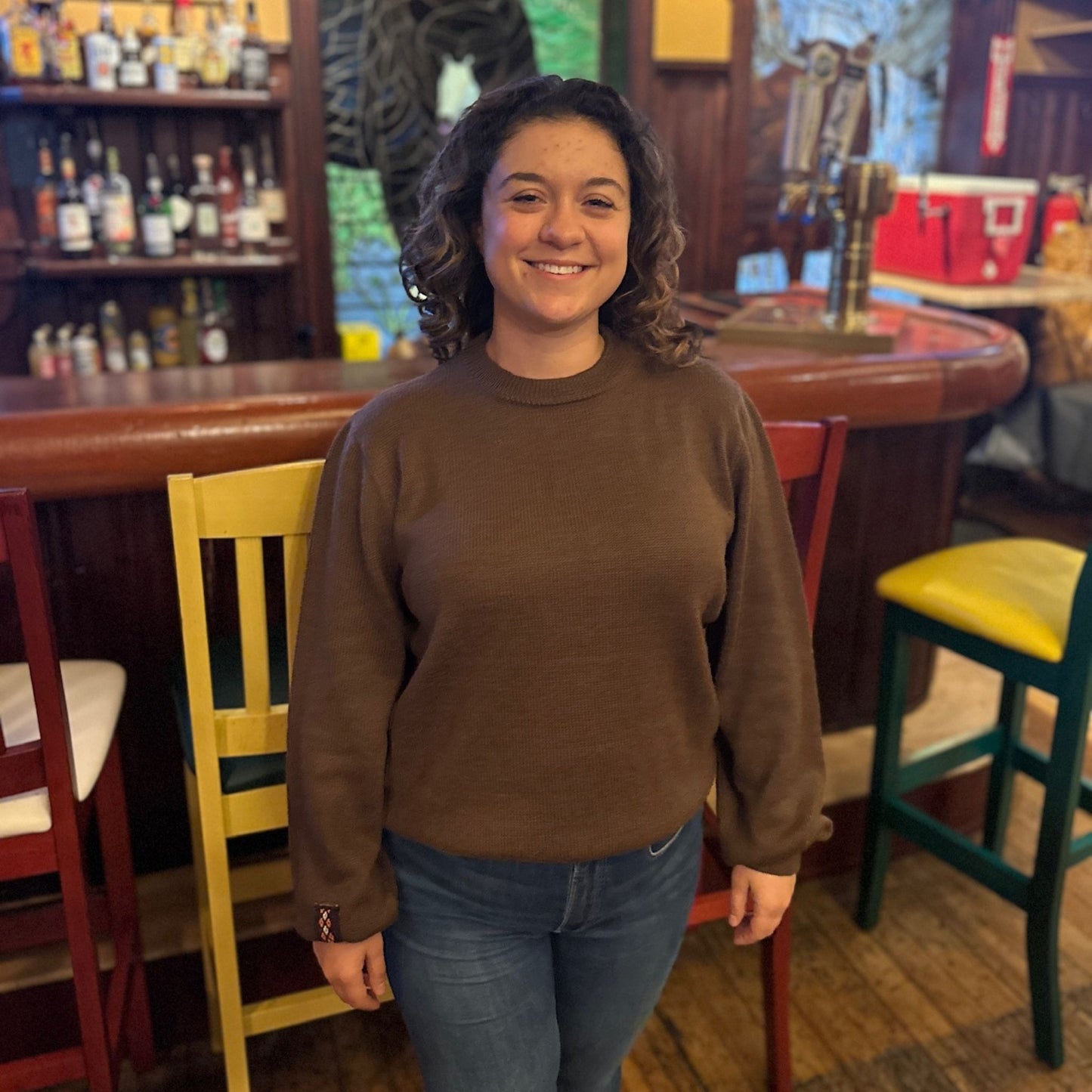 Person wearing a brown sweater standing in a bar with colorful chairs and shelves.