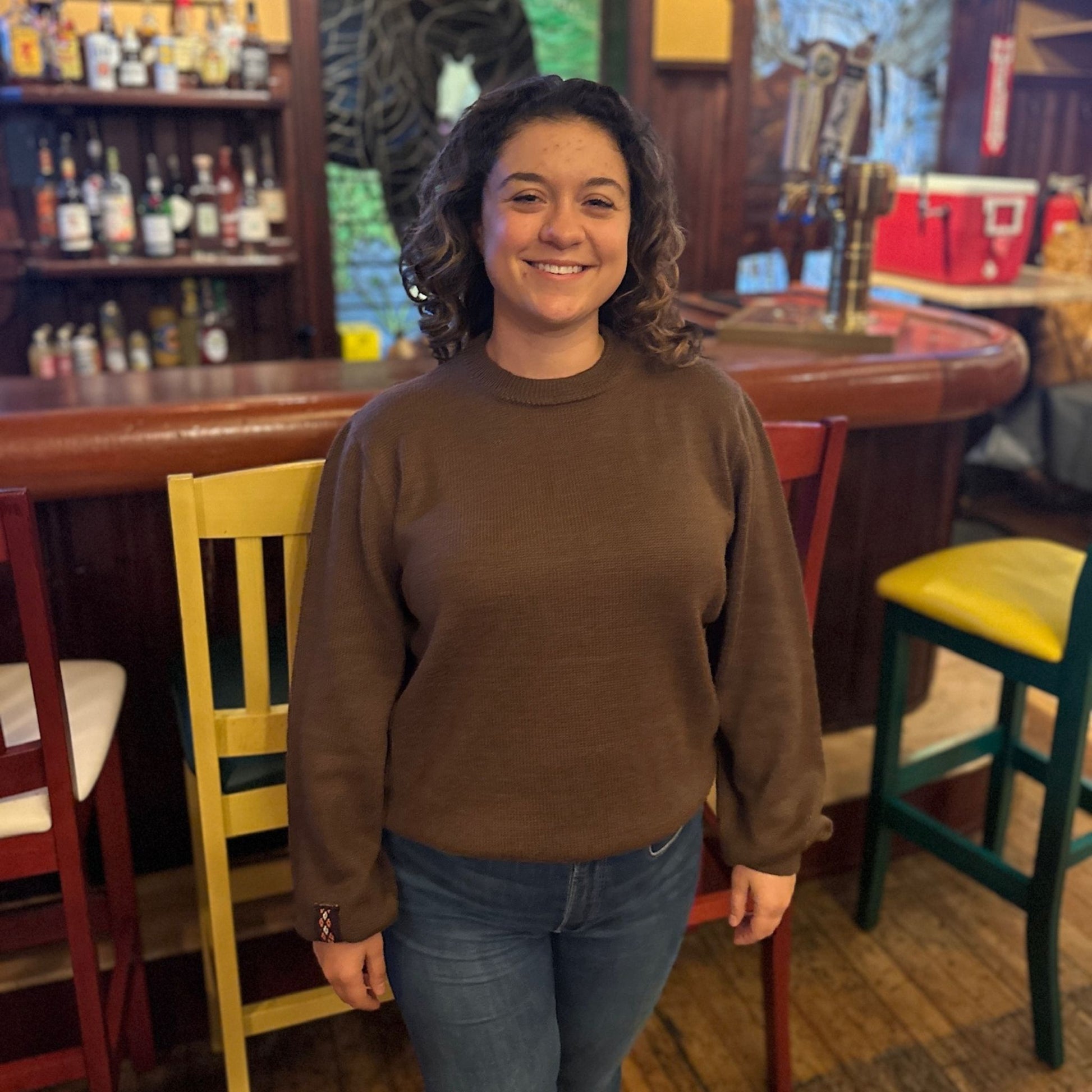 Person wearing a brown sweater standing in a bar with colorful chairs and shelves.