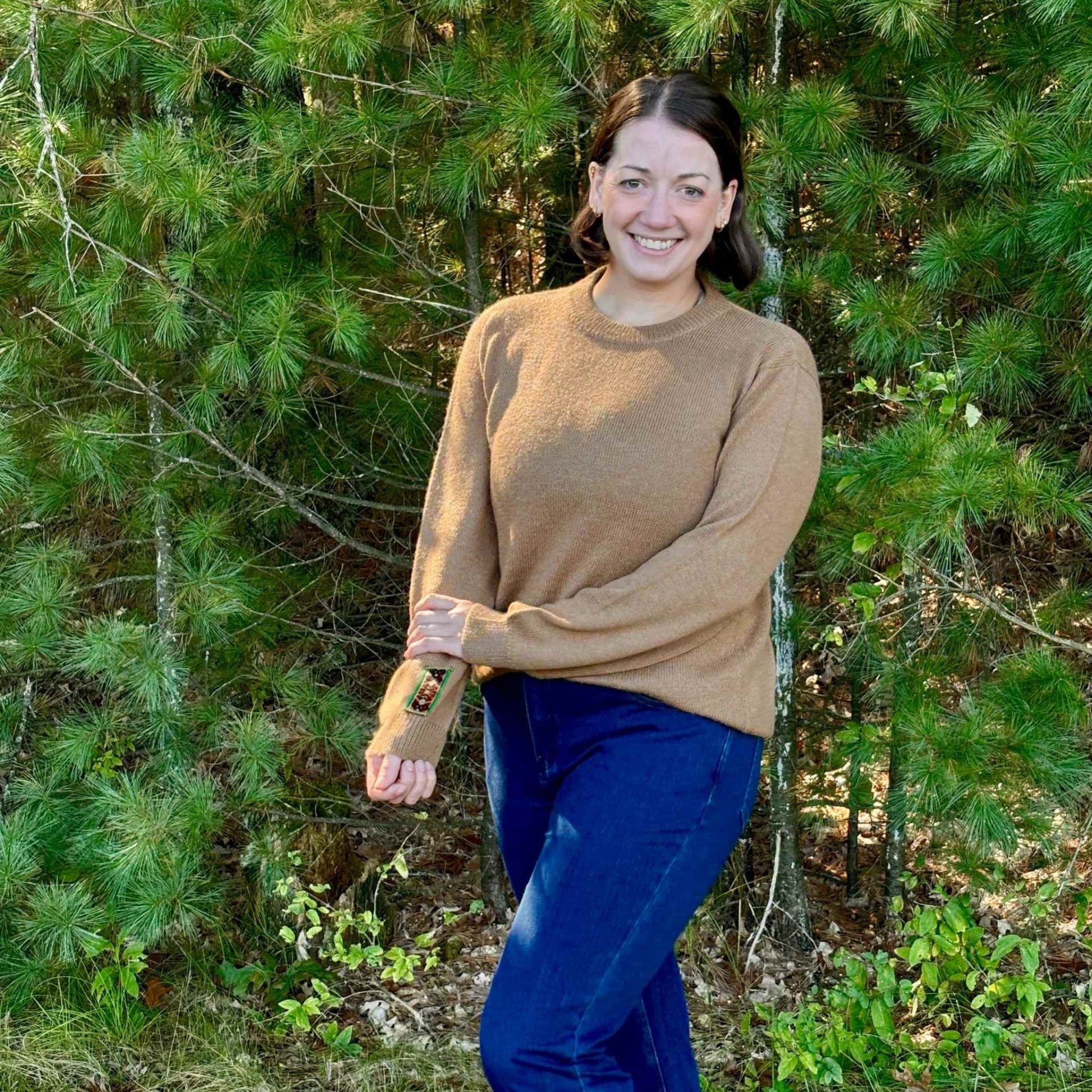 Woman in a hazelnut sweater and blue pants standing in a forest