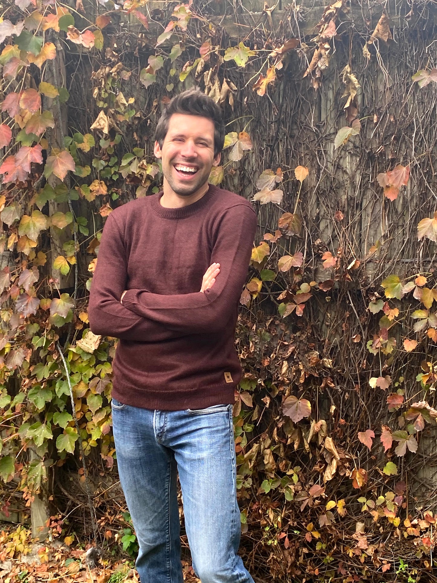 Man standing in front of a leafy wall, wearing a maroon sweater and blue jeans.