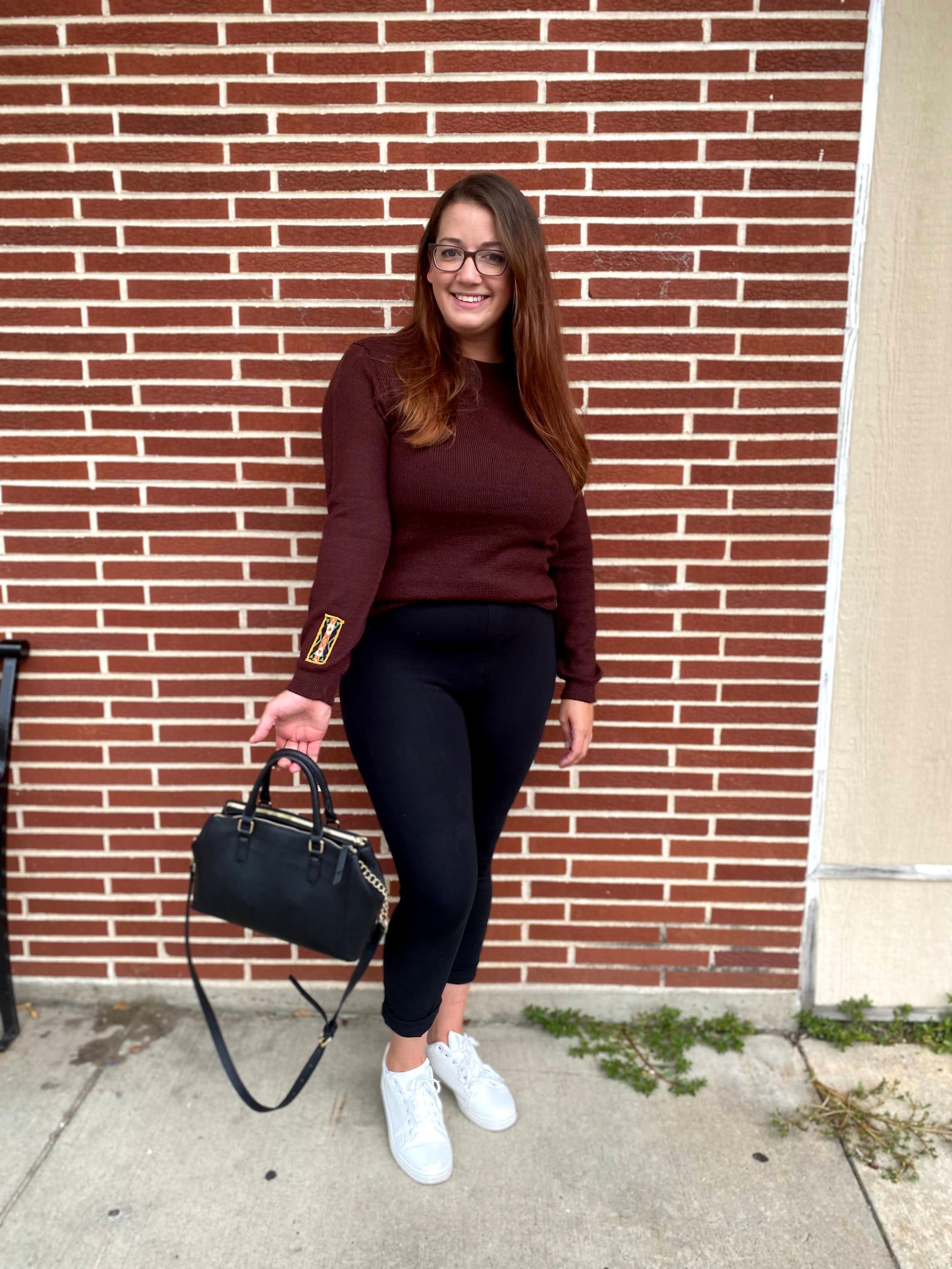 Woman in a maroon sweater and black pants standing against a brick wall.