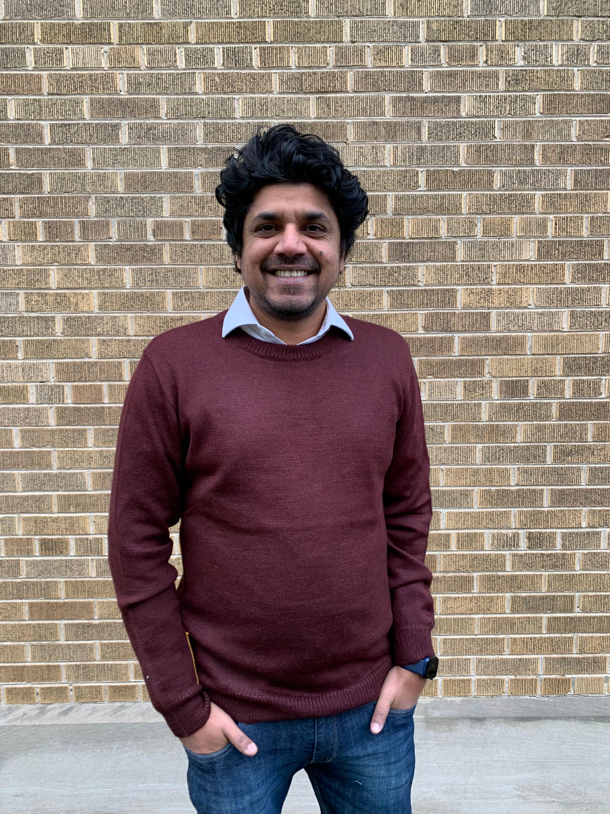 Man wearing a maroon sweater and blue jeans standing in front of a brick wall.