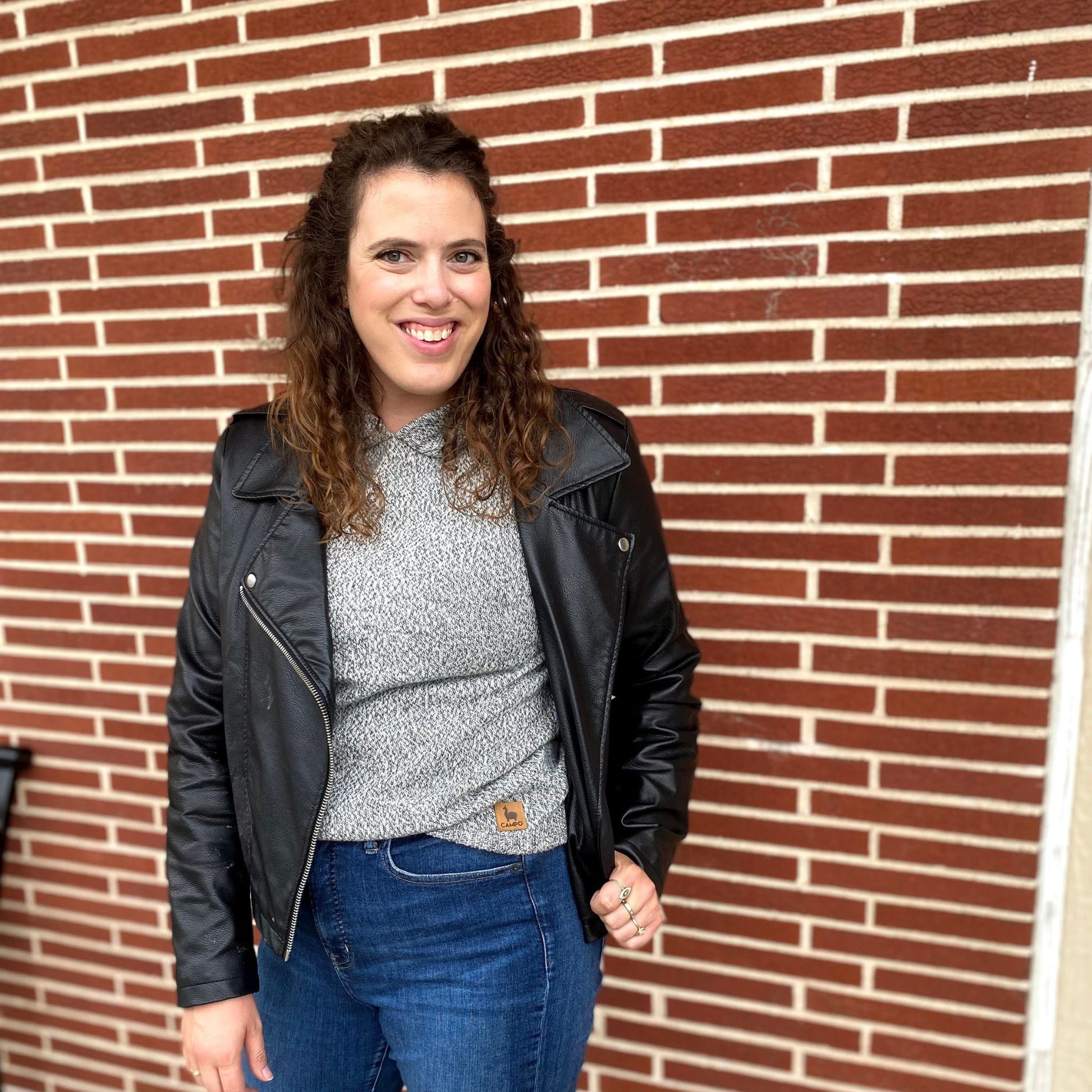 Woman wearing a black leather jacket, salt and peppre sweater, and blue jeans standing in front of a brick wall.