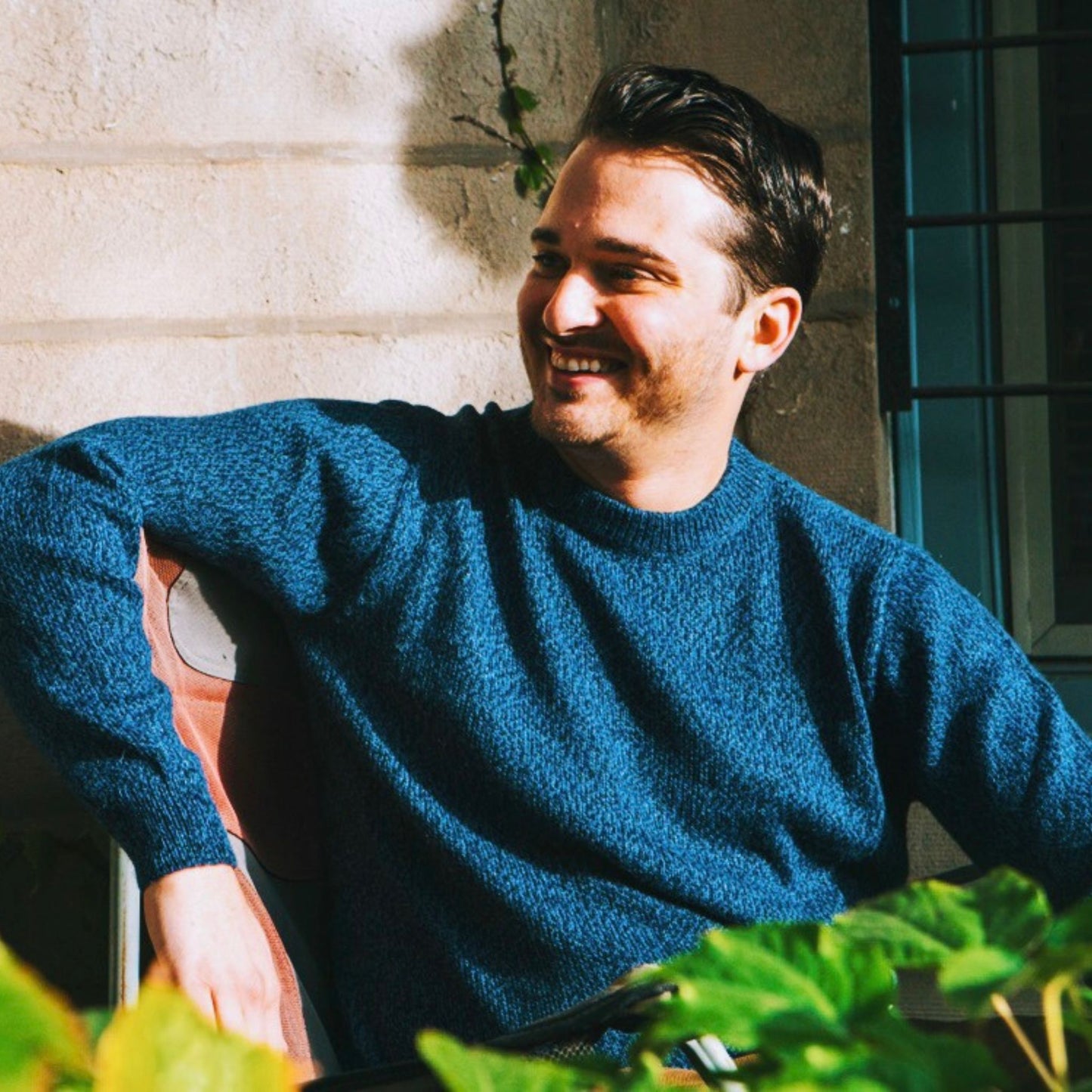 Man in a blue sweater sitting outdoors with plants and a building in the background