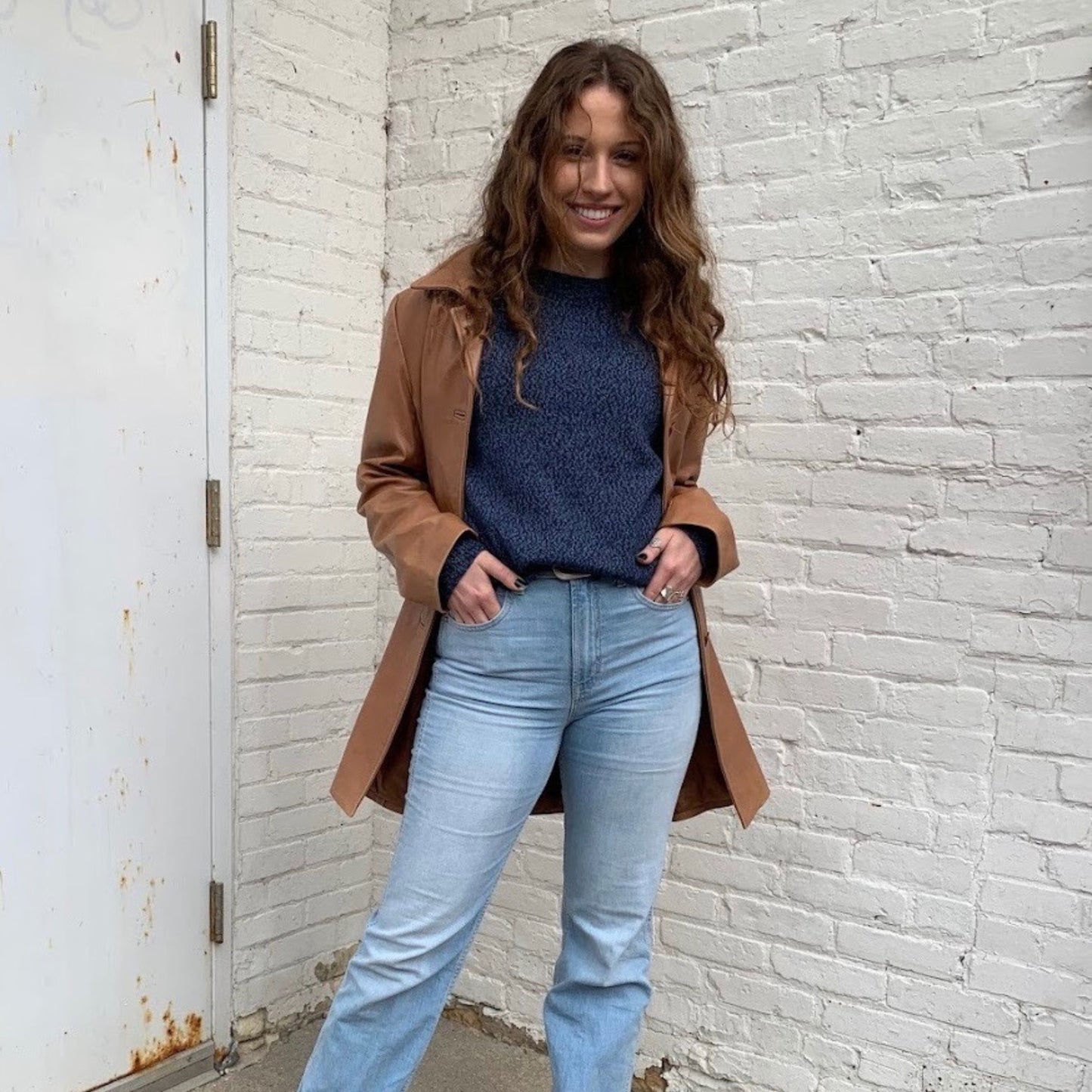 Woman wearing a brown coat, blue sweater, and light blue jeans standing against a white brick wall.