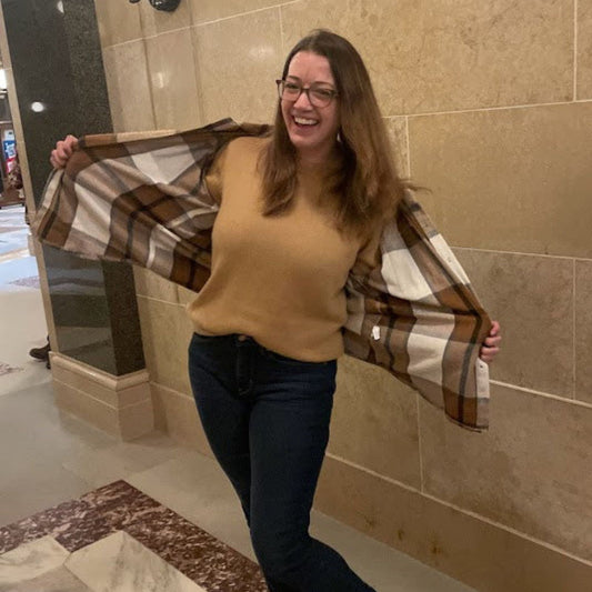 Woman wearing camel sweater holding a plaid shirt in a tiled room