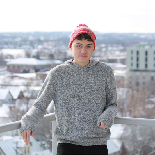 Person wearing a salt and pepper hoodie and red beanie standing on a balcony with a snowy cityscape background