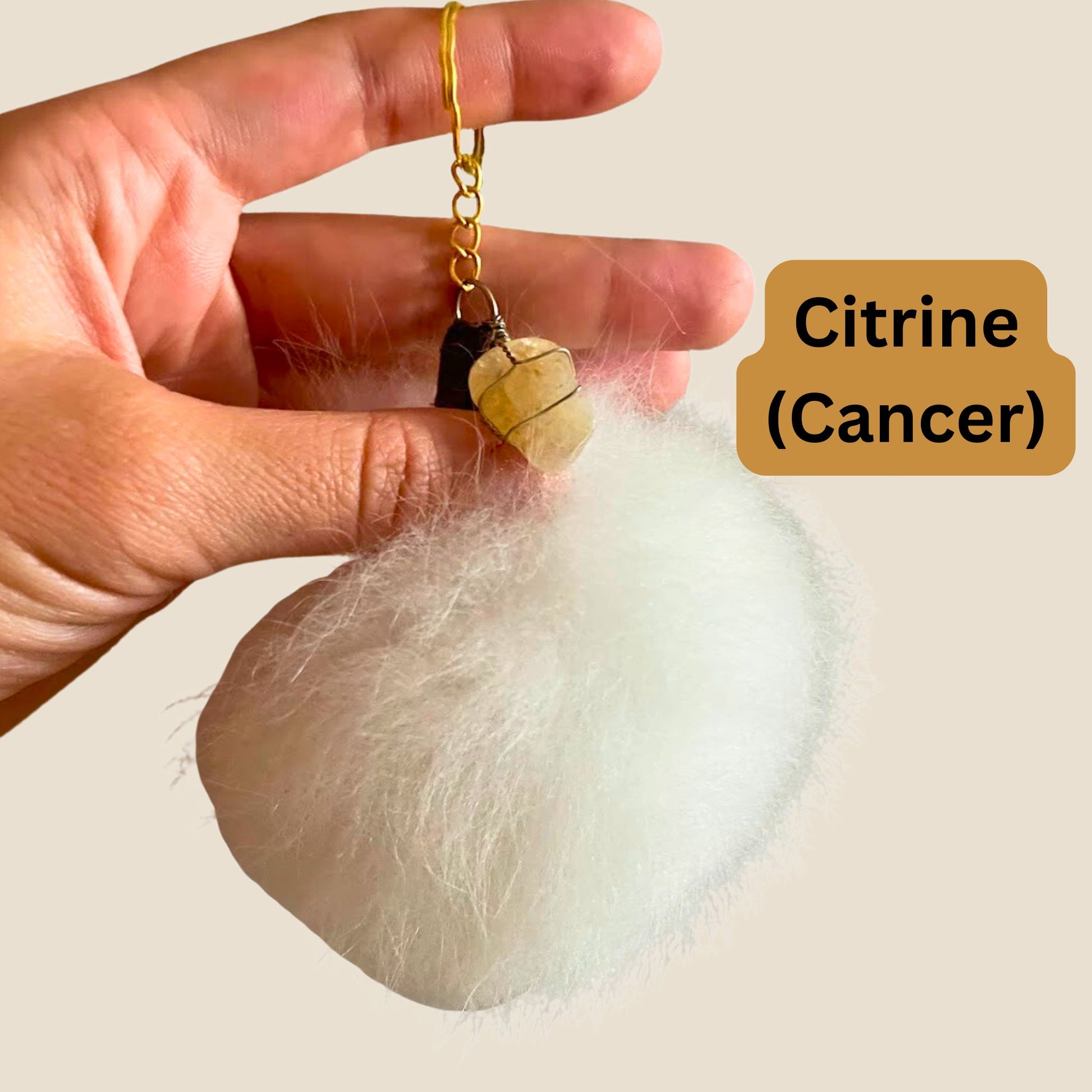 Hand holding a citrine stone keychain with a white fluffy object and 'Citrine (Cancer)' text on a beige background