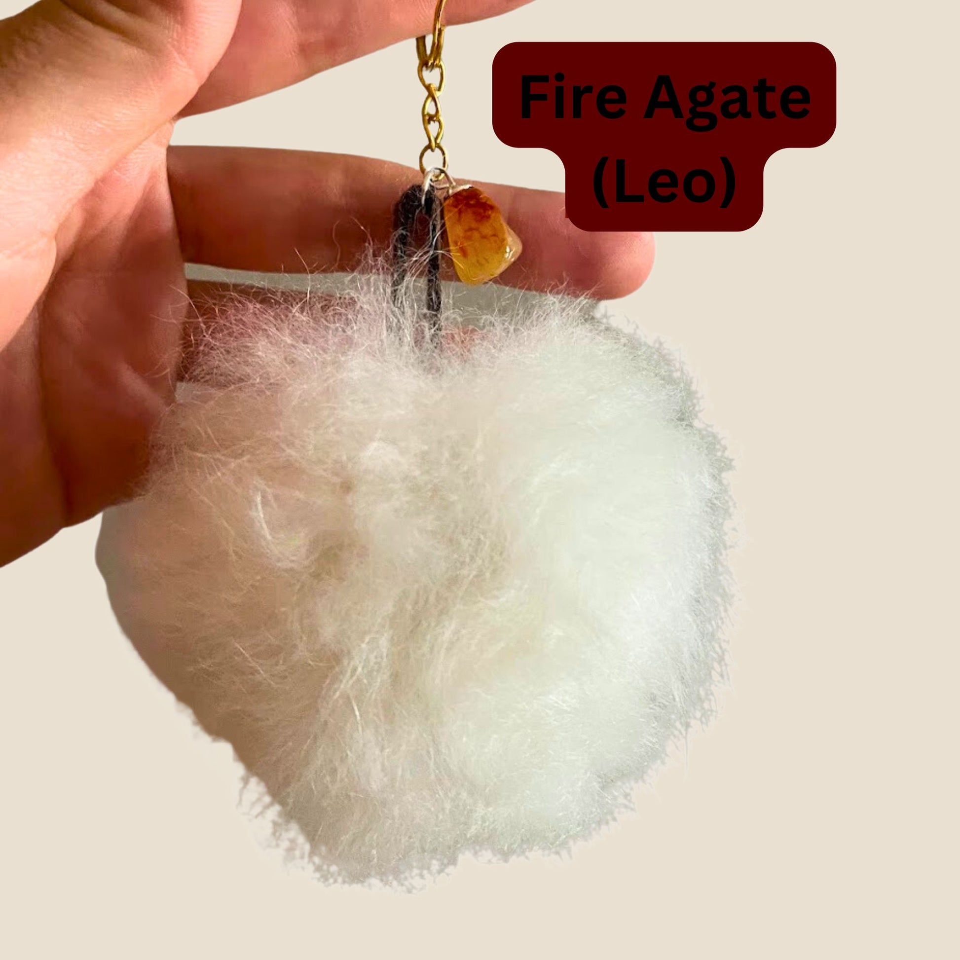 Fluffy keychain with a small yellow stone on a gold chain, held by a hand against a beige background.