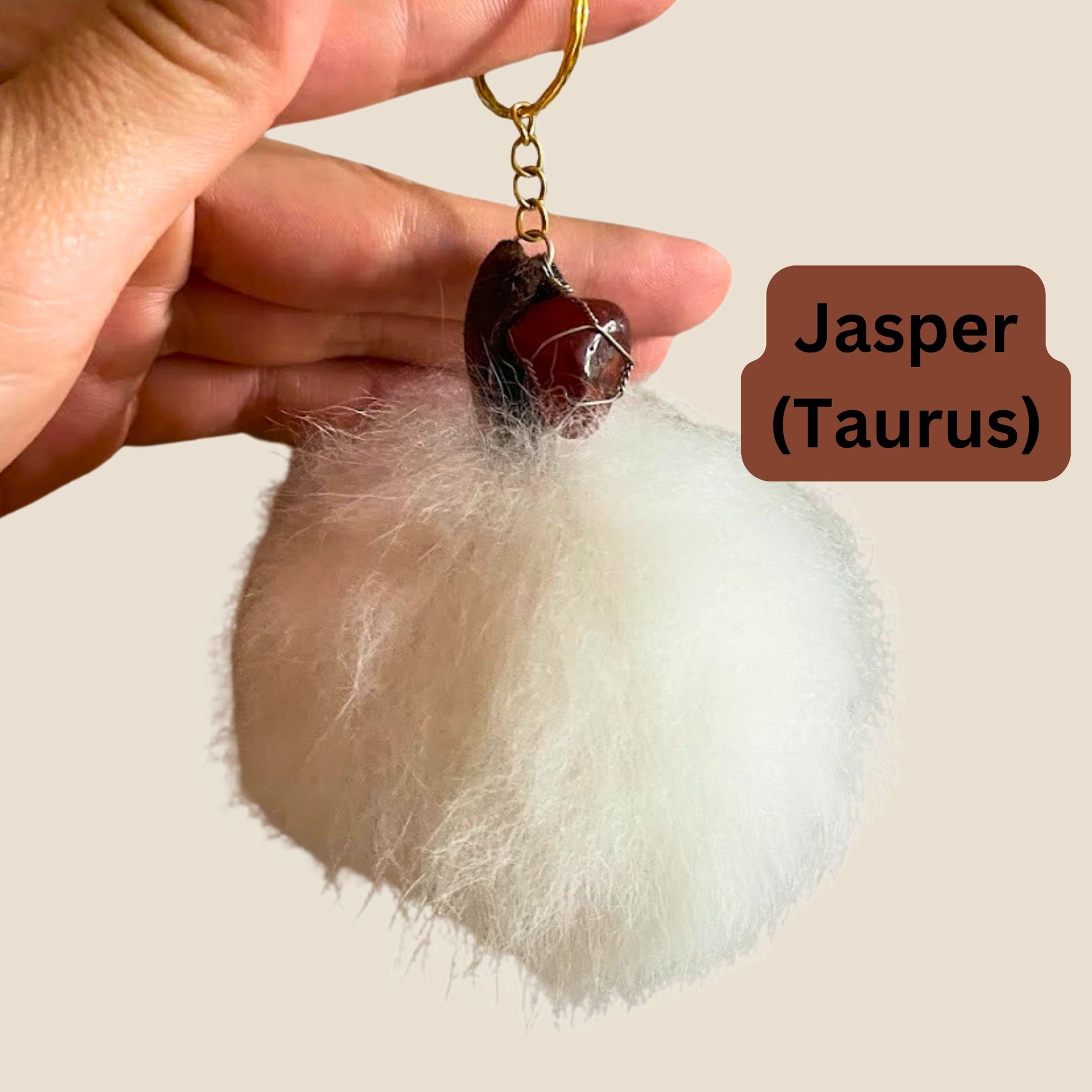 Fluffy white keychain with a brown stone and gold chain, held by a hand against a plain background.