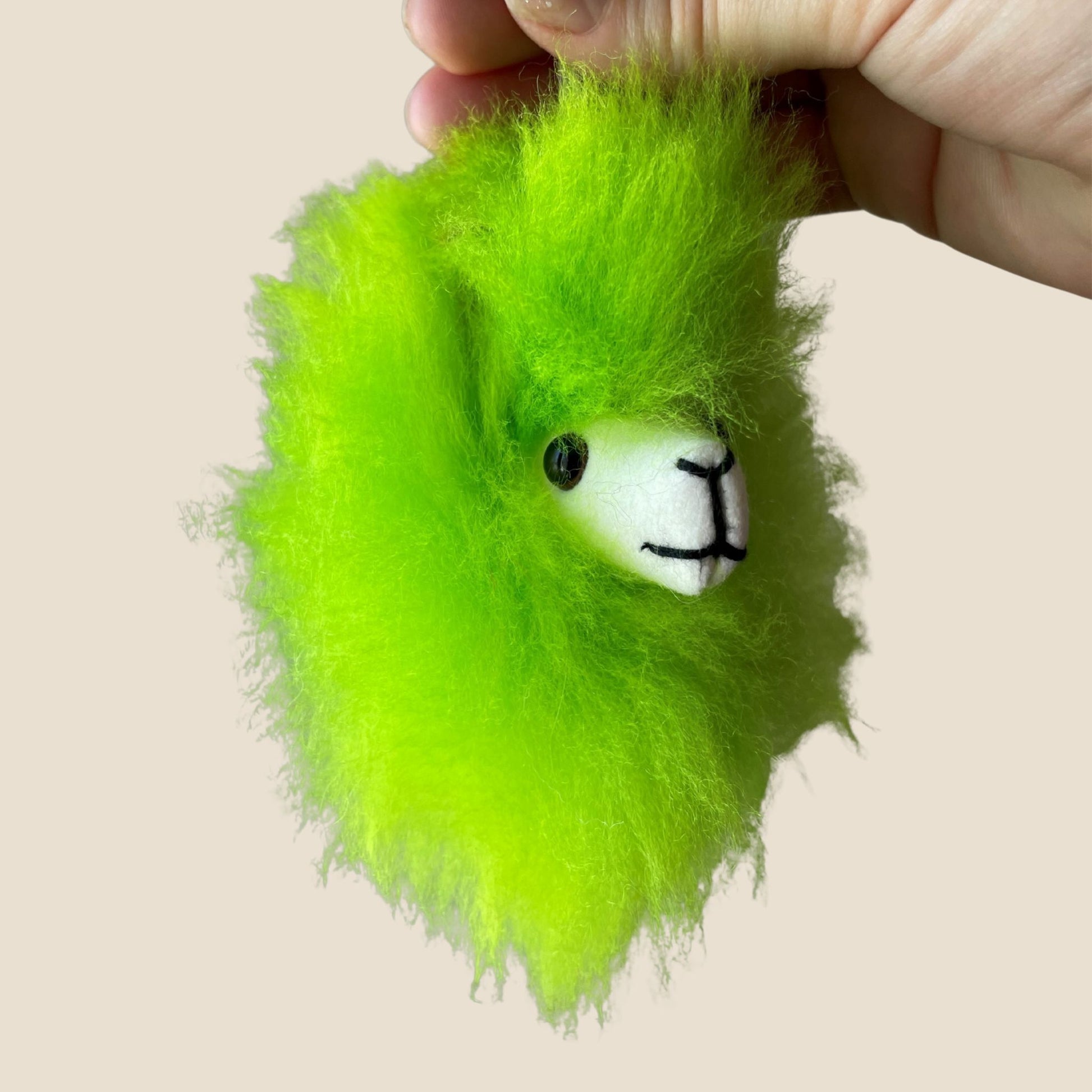 Green fluffy toy with a white face and black eyes held by a hand on a beige background