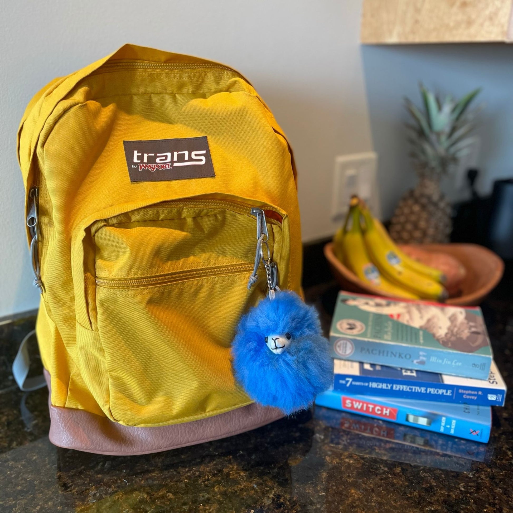 Yellow backpack with a blue pom-pom keychain on a surface with books and bananas in the background.