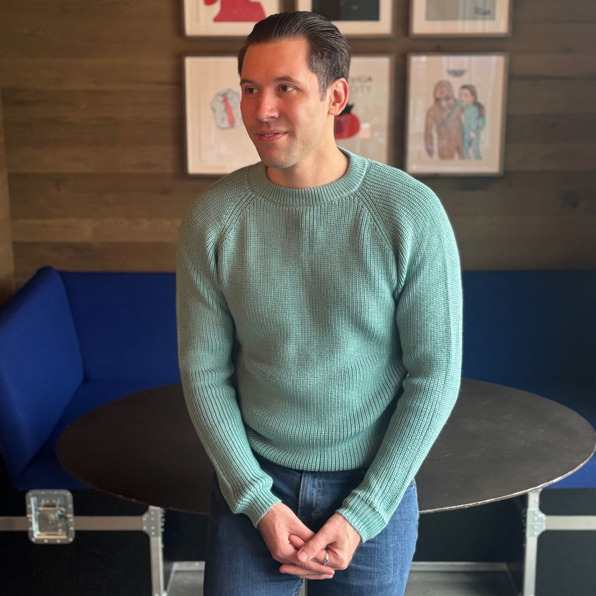 Man wearing a teal sweater standing in a room with blue couch and framed pictures on the wall.