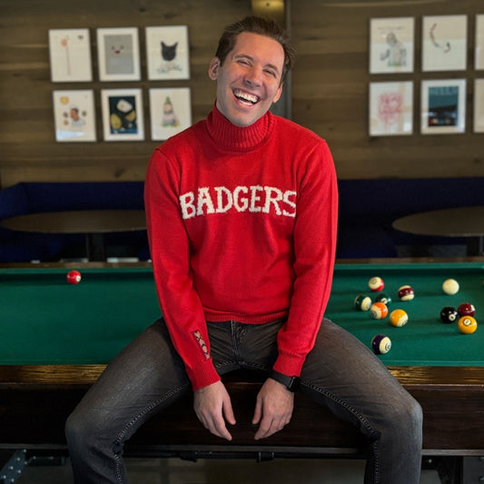 Man wearing a red sweater with 'BADGERS' sitting on a pool table.