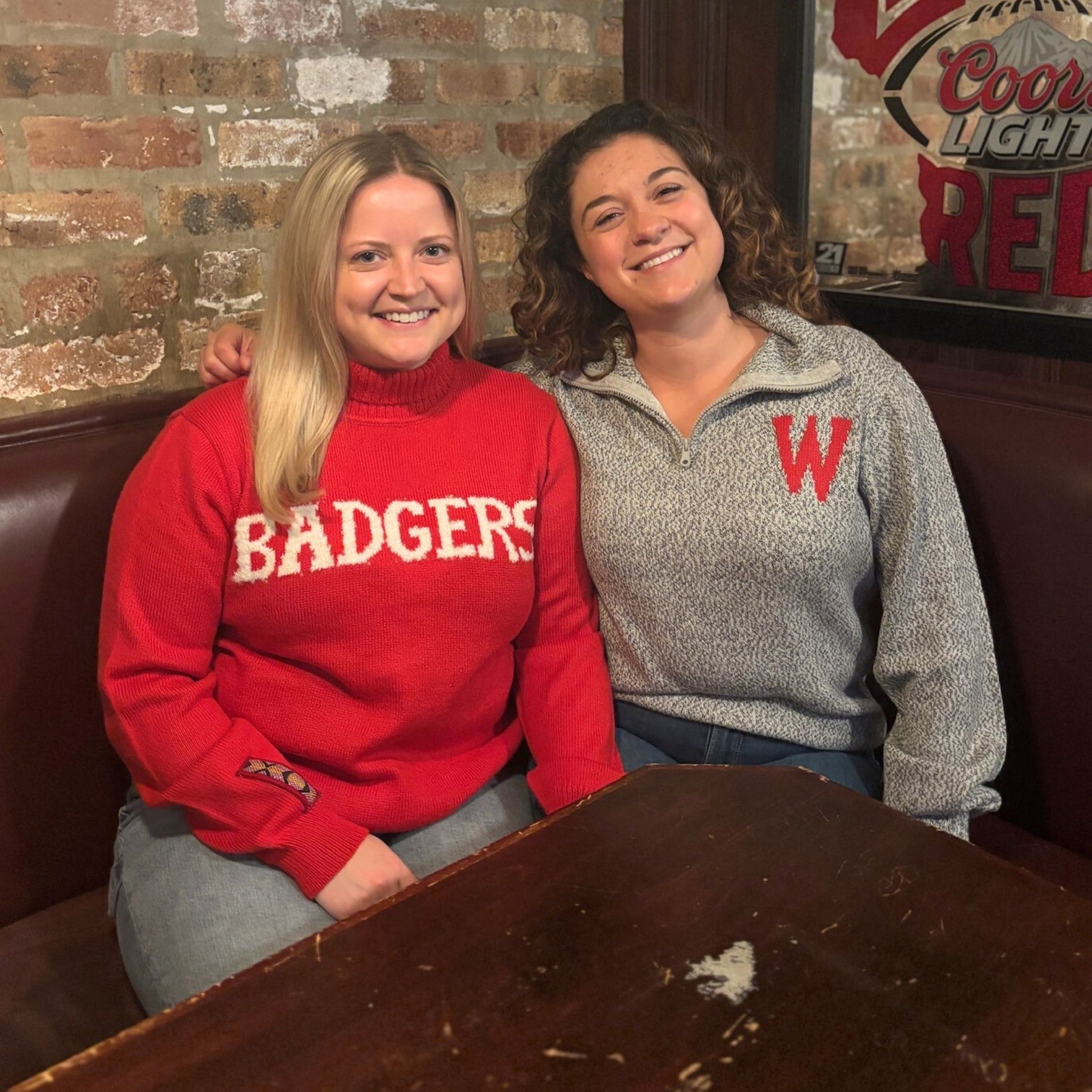 Two women sitting together in a casual setting with a brick wall and Coors Light sign in the background.