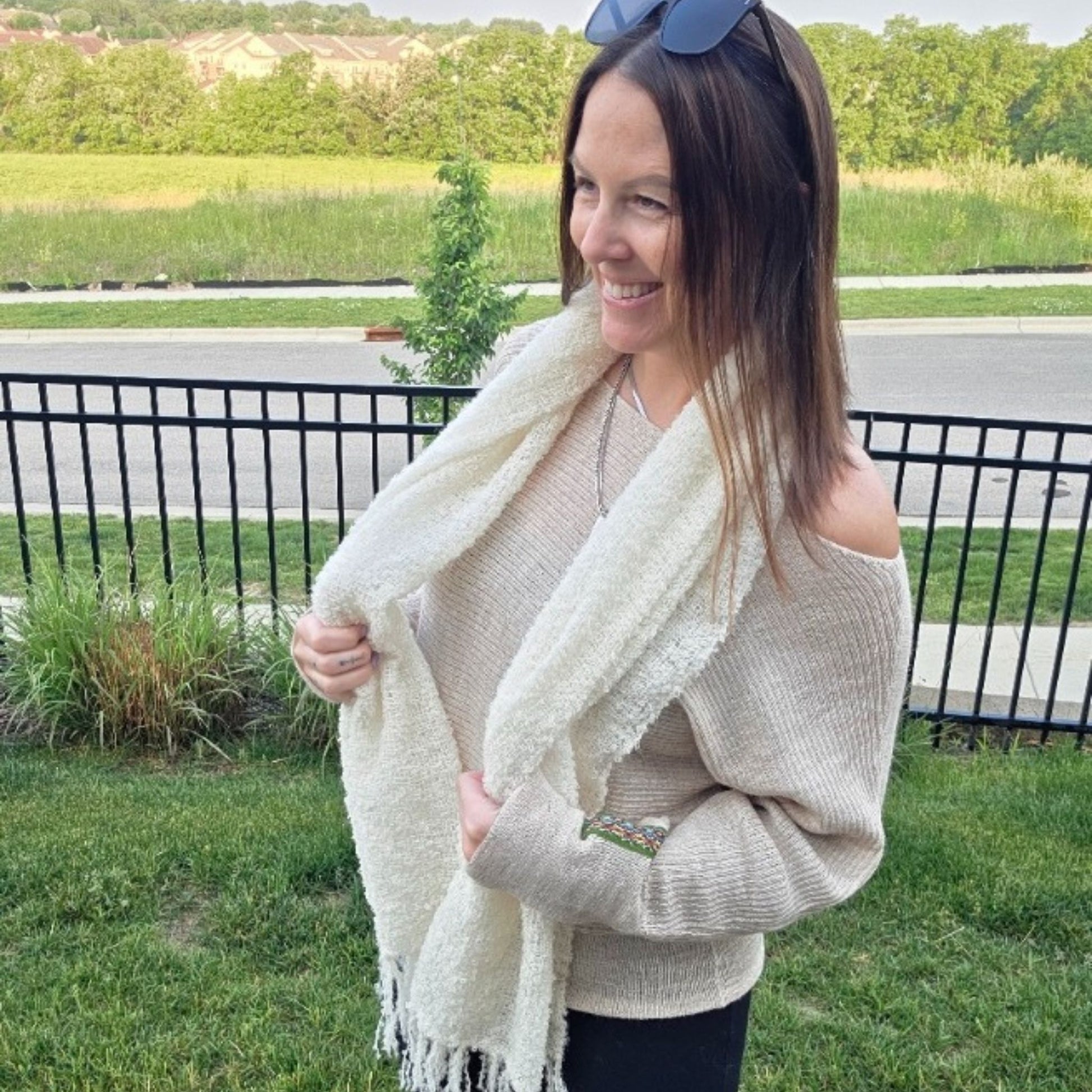 Woman wearing a beige sweater and white scarf outdoors with greenery in the background