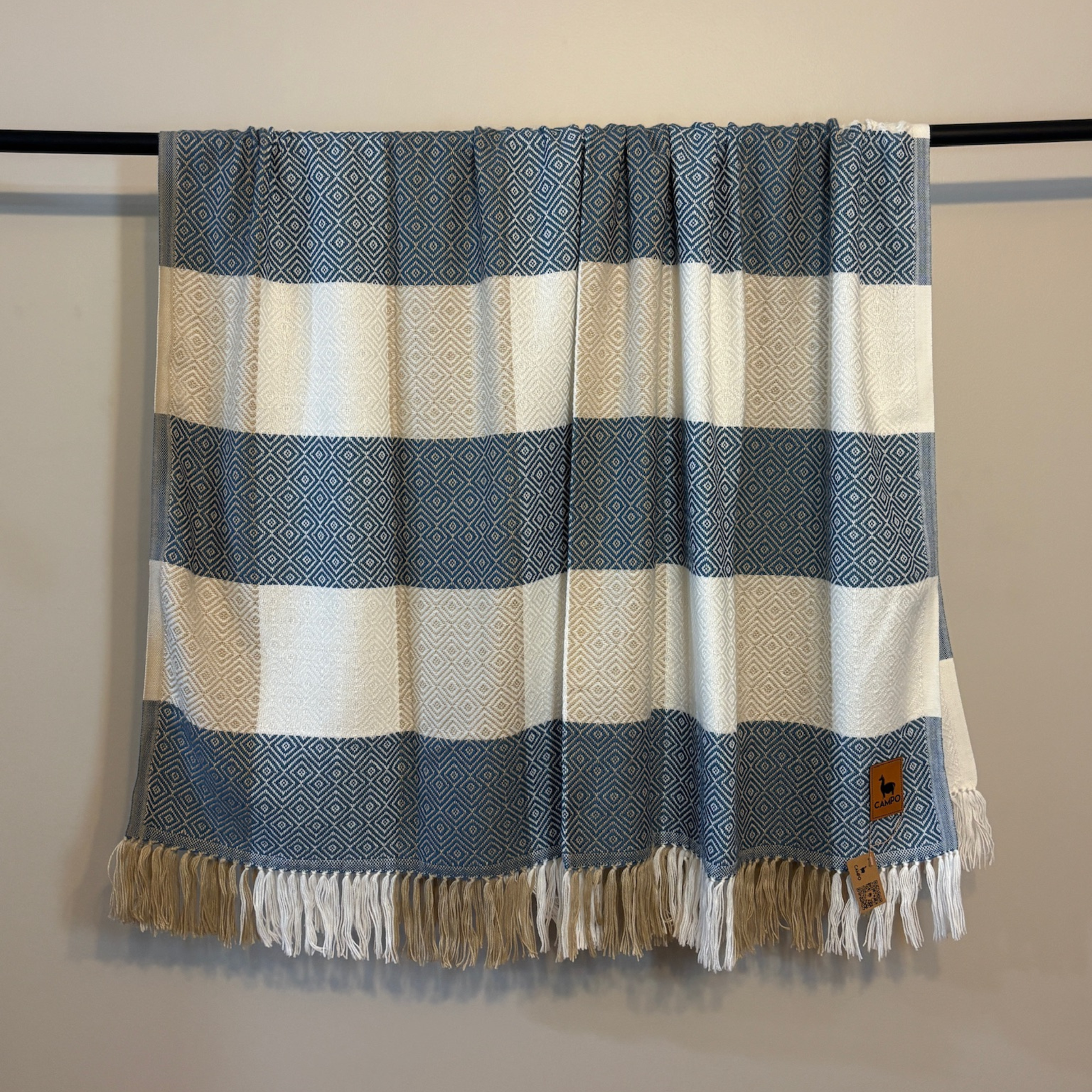 Striped blanket with blue and beige pattern draped over a black rod against a beige wall.