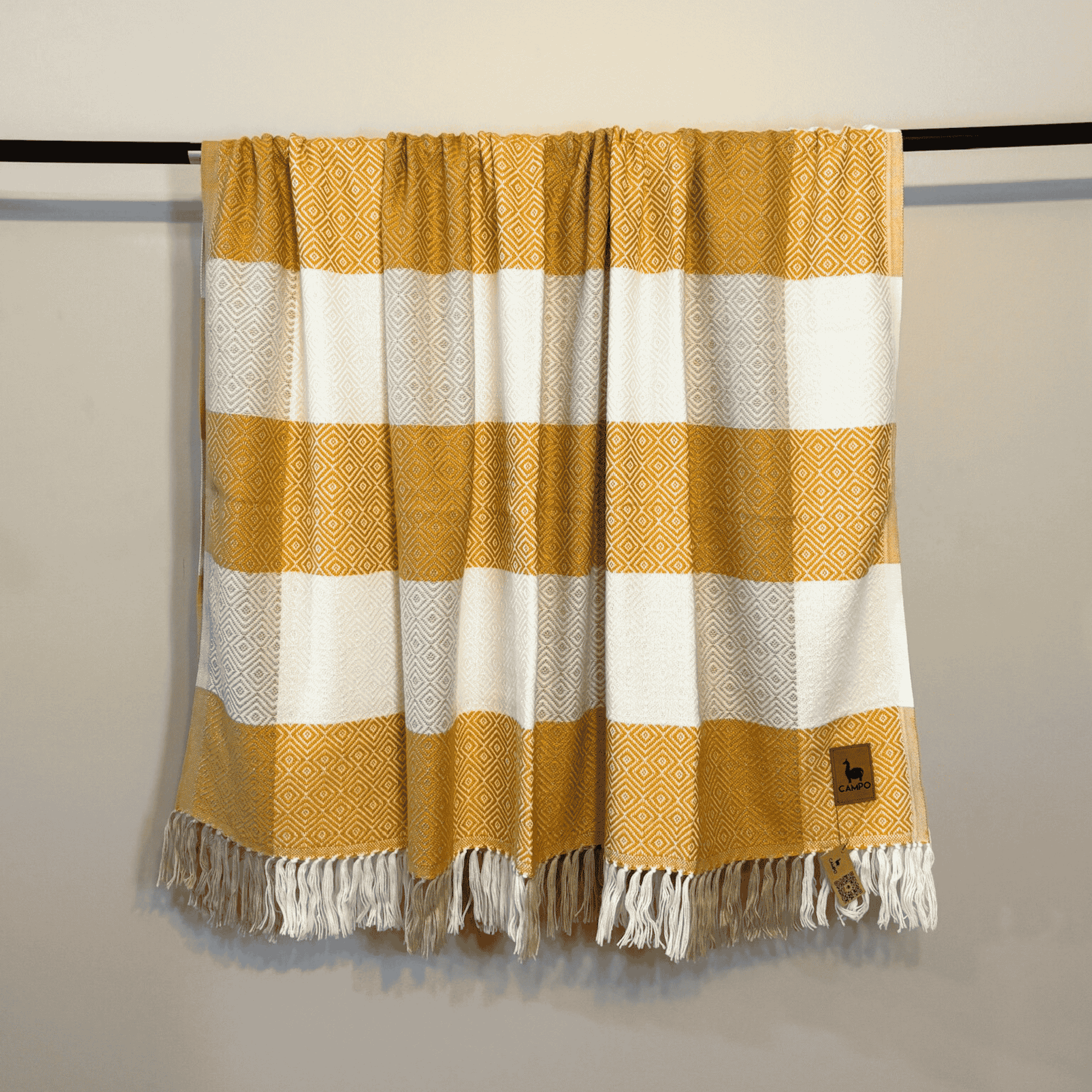 Mustard and white checkered blanket with fringes on a black rod against a beige wall.