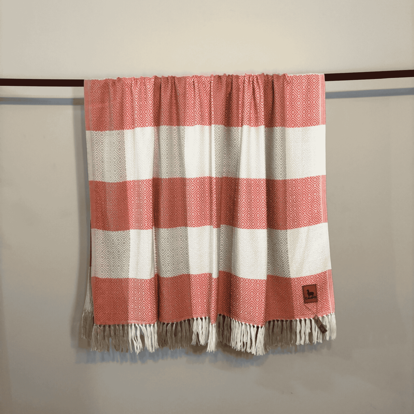 Rose and white striped blanket with fringes hanging on a rod against a beige wall.