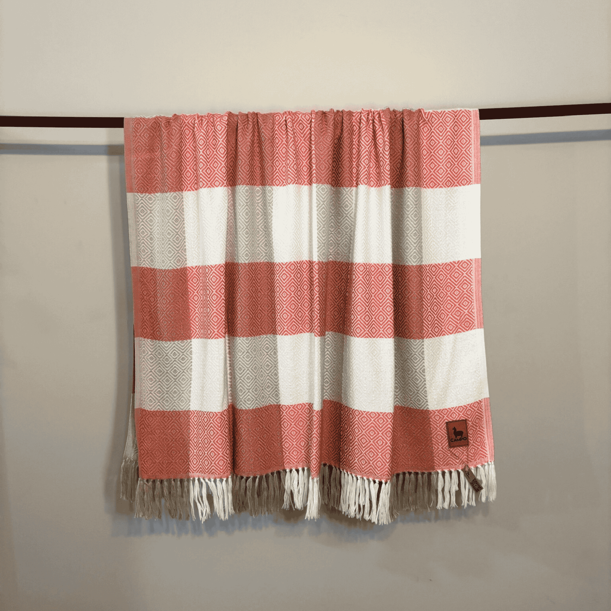 Rose and white striped blanket with fringes hanging on a rod against a beige wall.