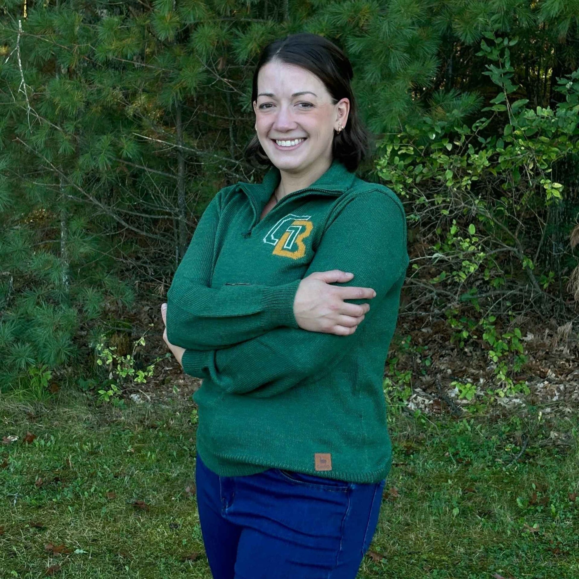 Woman wearing a green sweater with a logo standing in a forested area