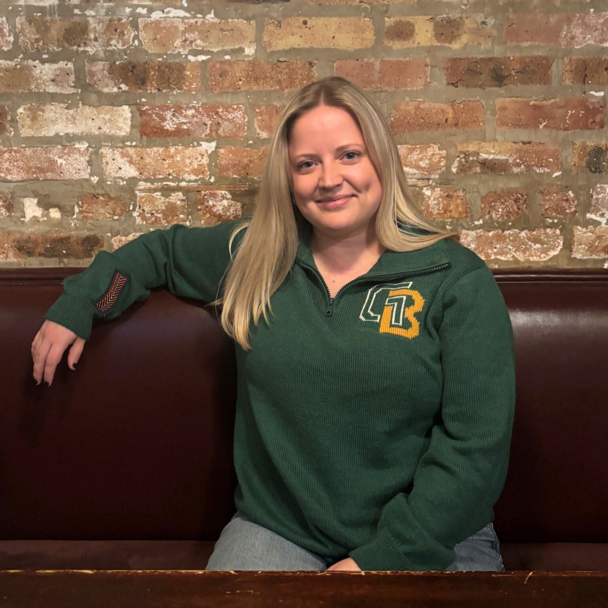 Woman wearing a green sweater with a logo, sitting on a brown leather couch against a brick wall.