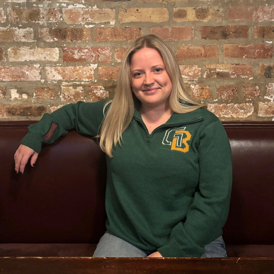 Woman wearing a green sweater with a logo, sitting on a brown leather couch against a brick wall.