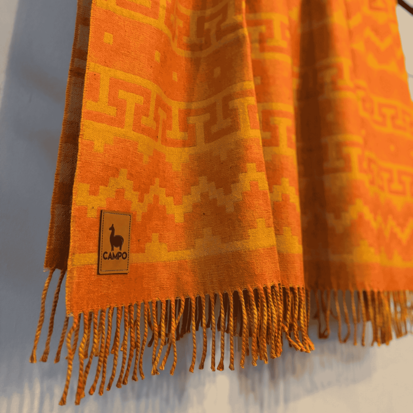 Orange patterned blanket with fringes and a brand logo on a neutral background