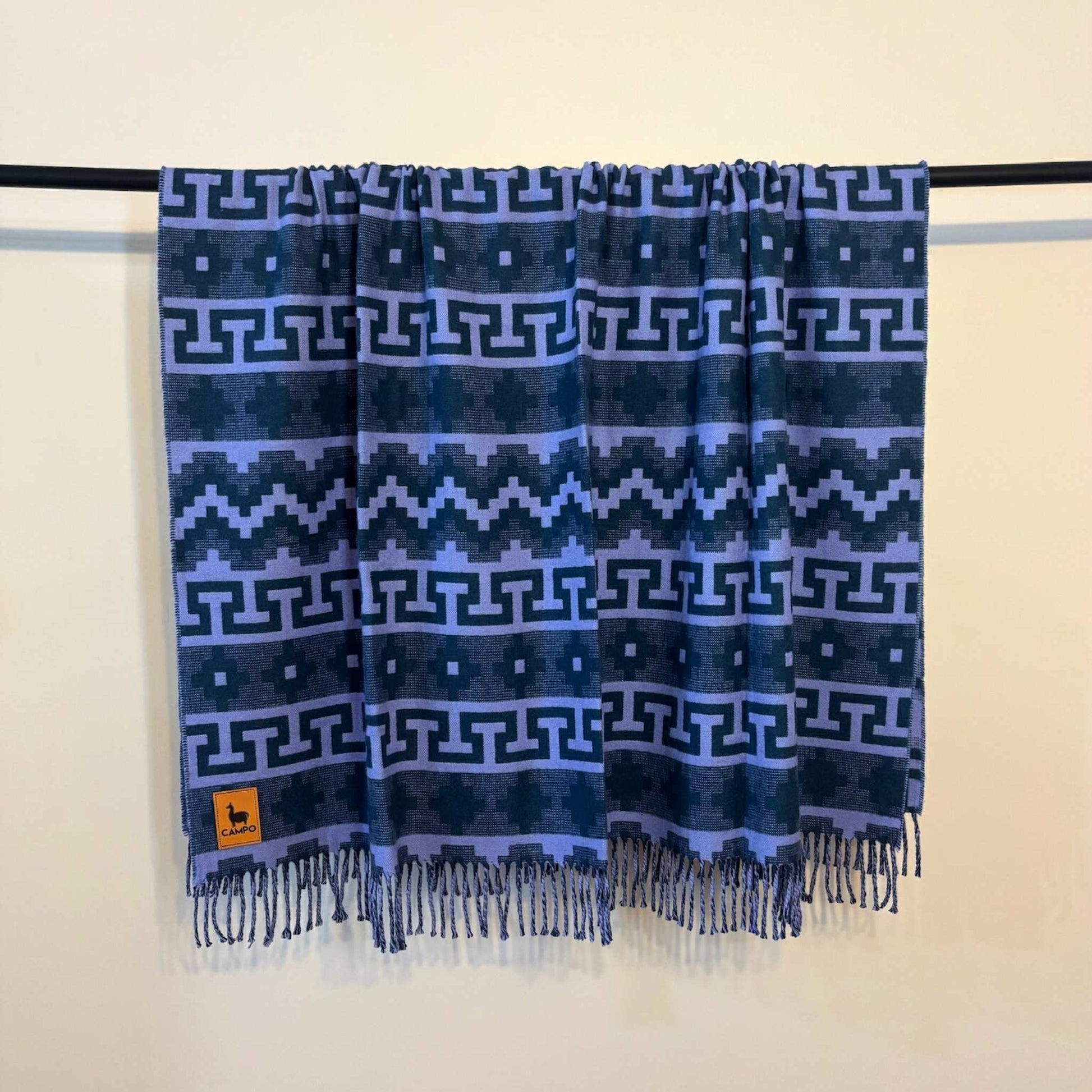Sapphire blue patterned scarf with fringes hanging on a black rod against a beige wall.