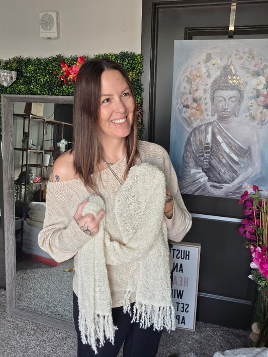Woman wearing a beige sweater with a white scarf, with a painting and flowers in the background.