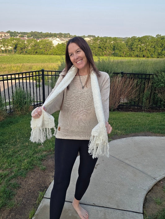 Woman wearing a beige sweater and white scarf standing on a sidewalk with grass and trees in the background