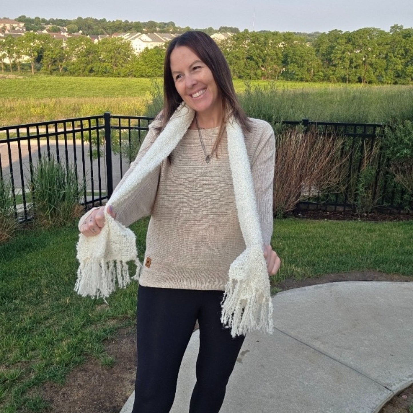 Woman wearing a beige sweater and white scarf outdoors with greenery in the background