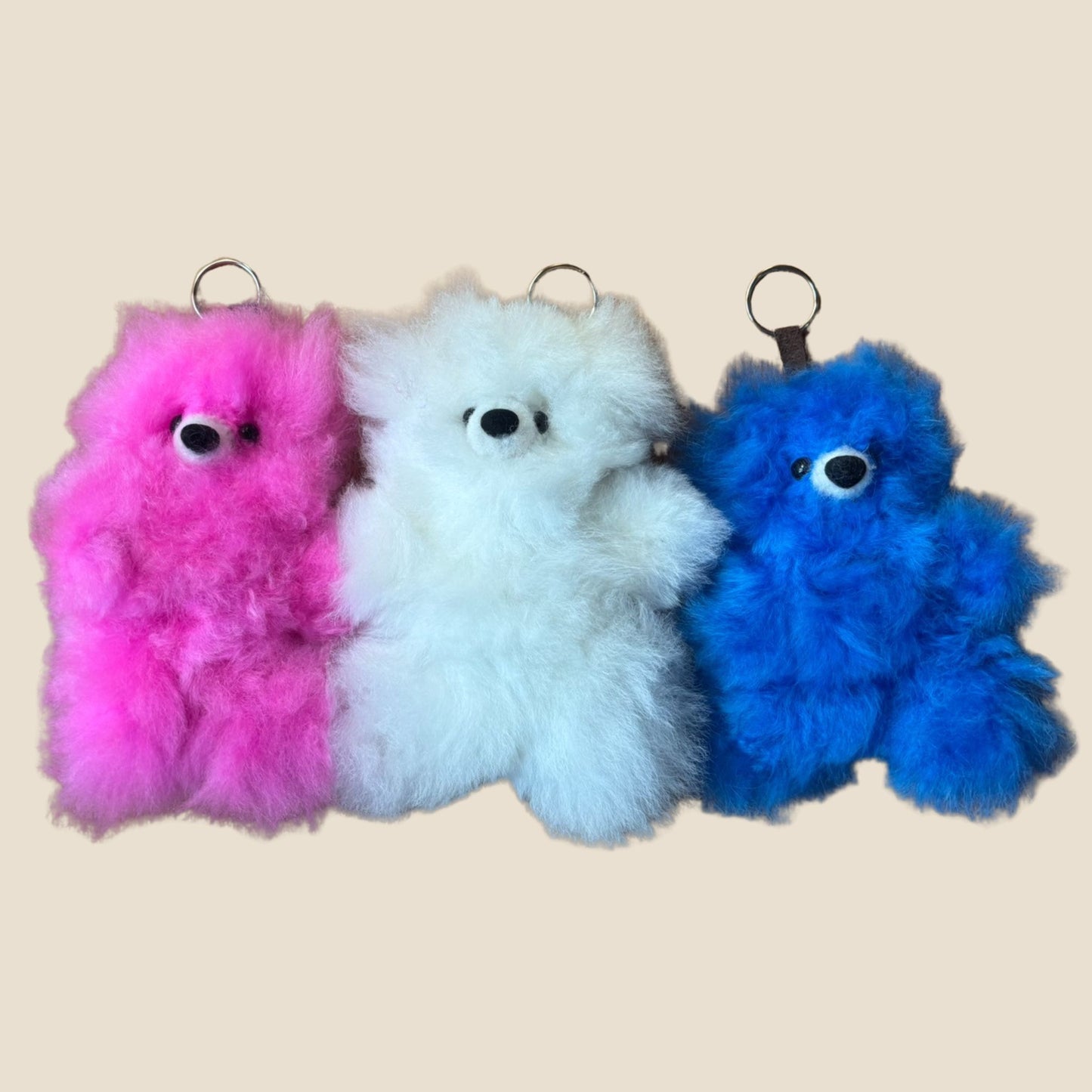 Three fluffy keychains in pink, white, and blue on a beige background