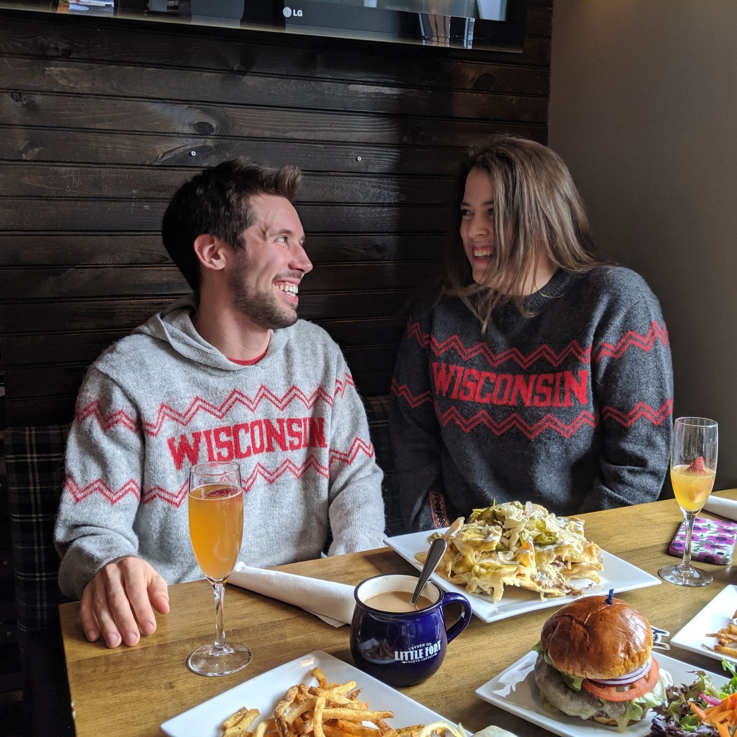 Two people wearing 'Wisconsin' sweaters sitting at a table with food and drinks.