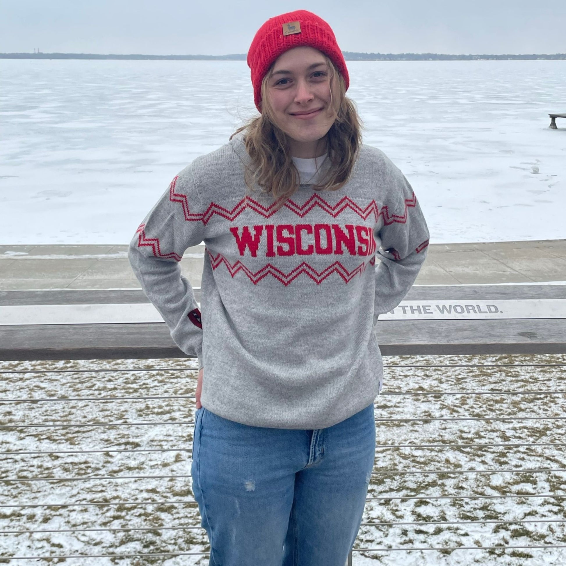 Person wearing a gray 'Wisconsin' sweater with a red beanie and jeans, standing outdoors in winter.
