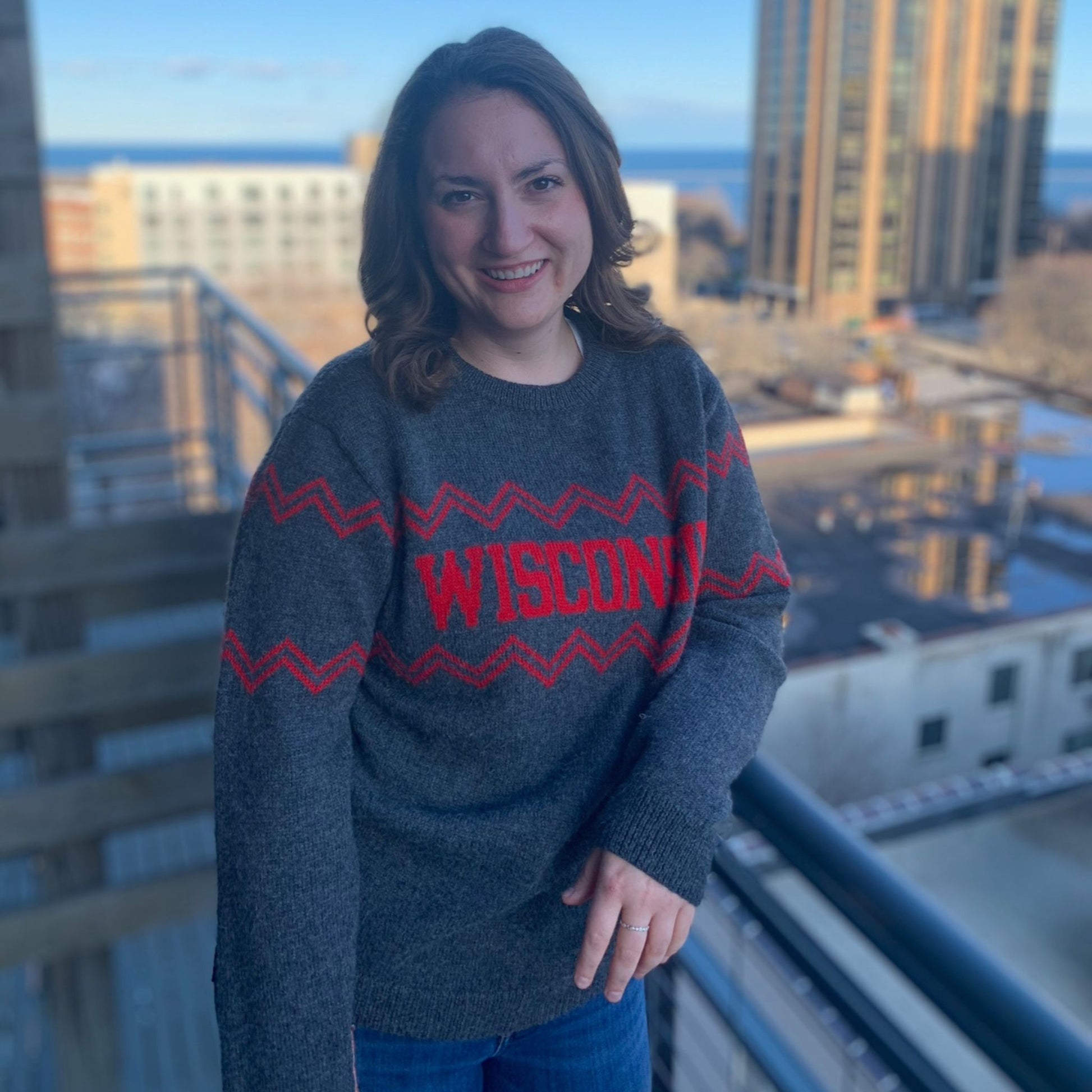 Person wearing a gray sweater with 'WISCONSIN' in red text, standing on a balcony with cityscape background.